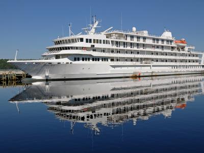 A large white cruise ship named "Pearl Mist" is docked at a wooden pier in calm water with a forested shoreline in the background.