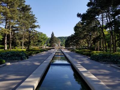 A long, rectangular reflecting pool or watercourse flanked by paved walkways and trees, leading towards a fountain structure in the distance.