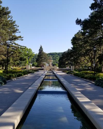 A long, rectangular reflecting pool or watercourse flanked by paved walkways and trees, leading towards a fountain structure in the distance.