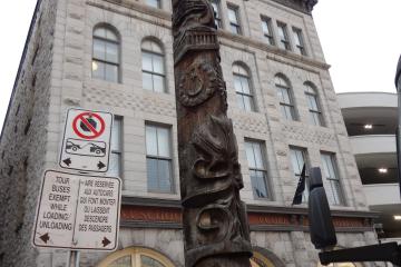 A carved wooden totem pole stands on a city sidewalk in front of a multi-story stone building with arched windows and storefronts, including signage for "Ottawa School of Art" and a bilingual bus loading sign.
