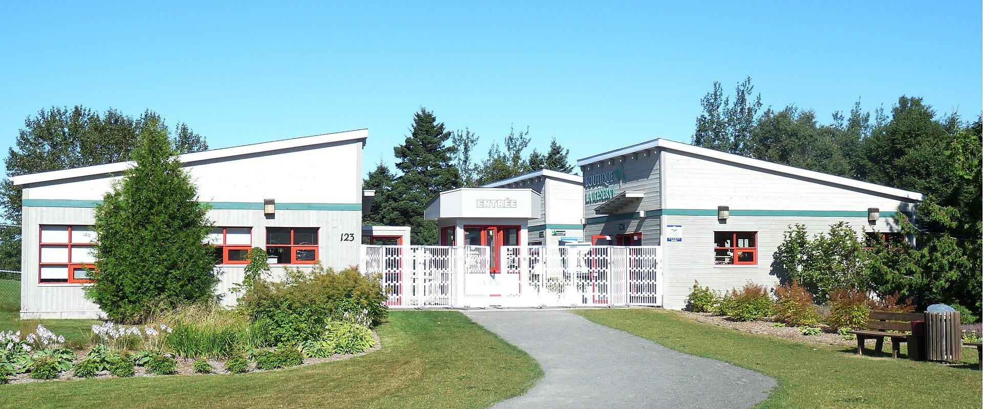 A single-story, modern-style building complex with white siding, green trim, and red window frames, featuring a central gated entrance labeled "ENTRANCE" and a sign on the right wing reading "BIOPARC DE LA GASPÉSIE".
