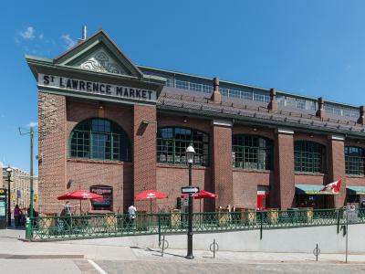 A low-angle exterior shot of a large red brick building with green trim and arched windows, featuring a prominent sign reading "ST LAWRENCE MARKET" on the facade, with outdoor seating under red umbrellas and a Canadian flag visible.