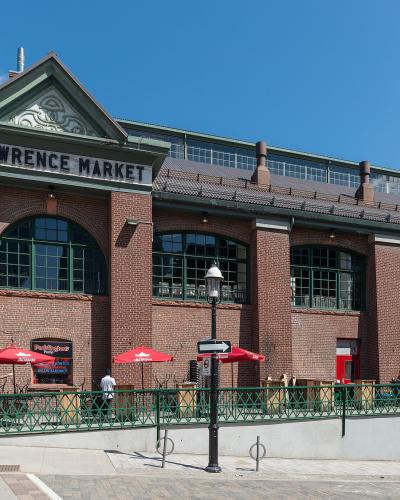 A low-angle exterior shot of a large red brick building with green trim and arched windows, featuring a prominent sign reading "ST LAWRENCE MARKET" on the facade, with outdoor seating under red umbrellas and a Canadian flag visible.