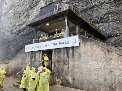 A concrete observation structure built into a rock face with visitors in yellow raincoats and a banner reading "JOURNEY BEHIND THE FALLS".