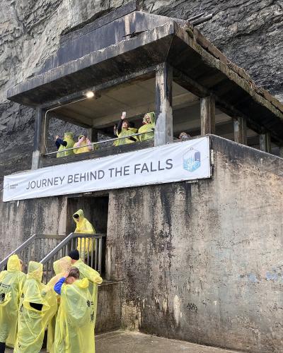 A concrete observation structure built into a rock face with visitors in yellow raincoats and a banner reading "JOURNEY BEHIND THE FALLS".