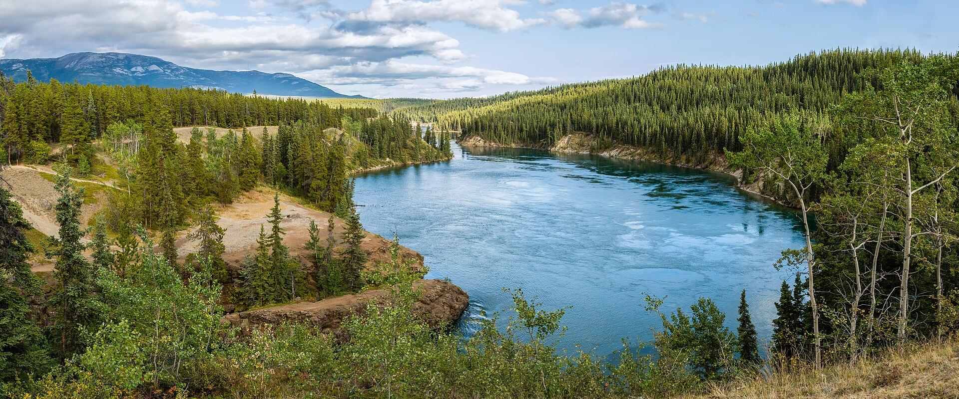 A panoramic landscape photograph showing a wide, blue river winding through a valley lined with dense coniferous forest and rolling hills under a cloudy sky.