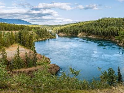 A panoramic landscape photograph showing a wide, blue river winding through a valley lined with dense coniferous forest and rolling hills under a cloudy sky.