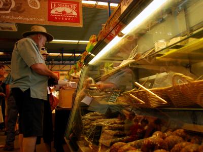 The image shows an indoor market stall with a customer browsing a refrigerated display case filled with sausages, under a red sign reading "LE MARCHÉ DU VIEUX-PORT QUÉBEC".