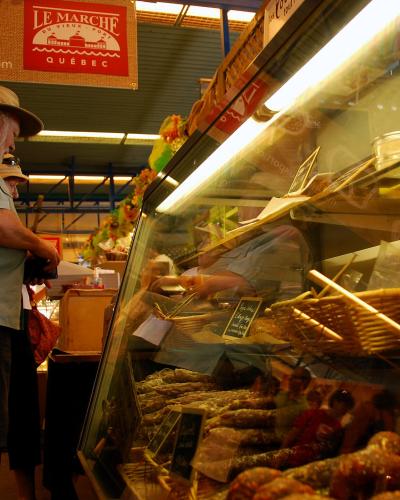 The image shows an indoor market stall with a customer browsing a refrigerated display case filled with sausages, under a red sign reading "LE MARCHÉ DU VIEUX-PORT QUÉBEC".