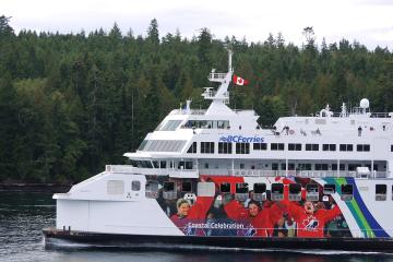 A large white BC Ferries vessel with a "Coastal Celebration" livery featuring athletes in red jackets, flying a Canadian flag, sailing past a forested coastline.