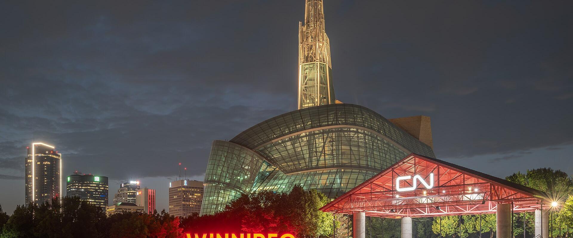 A night view of the Canadian Museum for Human Rights in Winnipeg, featuring its distinctive glass and stone architecture, a large illuminated "WINNIPEG" sign in the foreground, and a transit shelter with a "CN" logo.