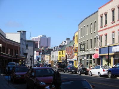 A street scene in an urban area with colorful historic buildings, parked cars, and a modern high-rise building in the background under a clear blue sky.