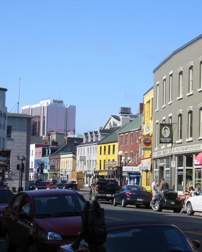 A street scene in an urban area with colorful historic buildings, parked cars, and a modern high-rise building in the background under a clear blue sky.