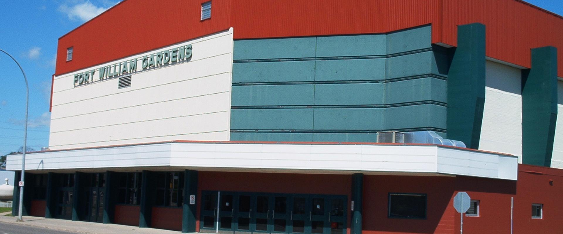 The image shows the exterior of a large entertainment arena building with red, white, and teal paneling under a blue sky, featuring prominent signage on the facade.