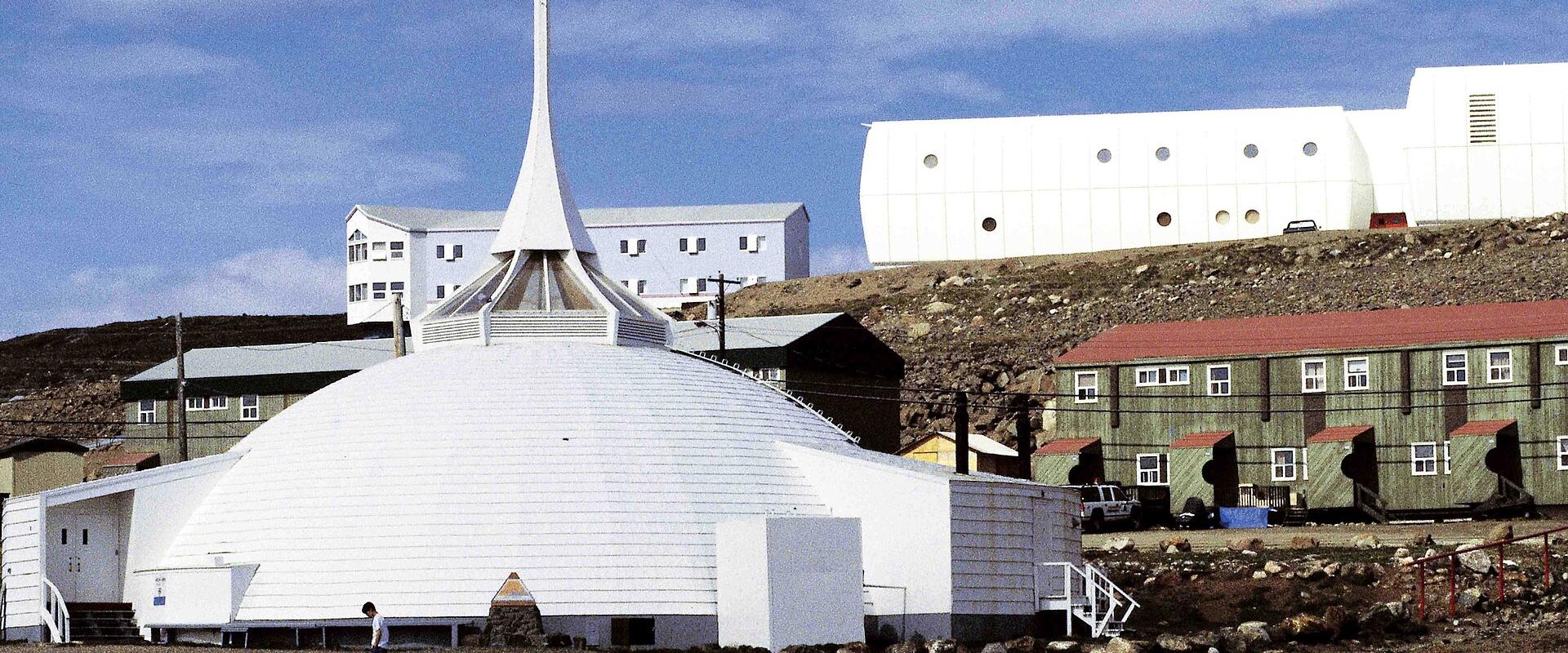 A white, dome-shaped church with a tall, slender spire topped by a cross, situated in a barren, rocky landscape with other buildings in the background.