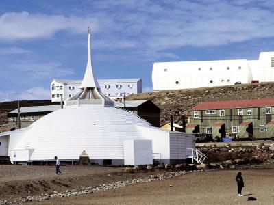 A white, dome-shaped church with a tall, slender spire topped by a cross, situated in a barren, rocky landscape with other buildings in the background.