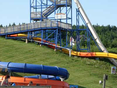 A tall blue steel tower structure supporting a steep white water slide and a multi-colored slide, with people waiting at the top and others near the bottom on a grassy hill.