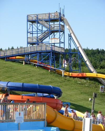 A tall blue steel tower structure supporting a steep white water slide and a multi-colored slide, with people waiting at the top and others near the bottom on a grassy hill.