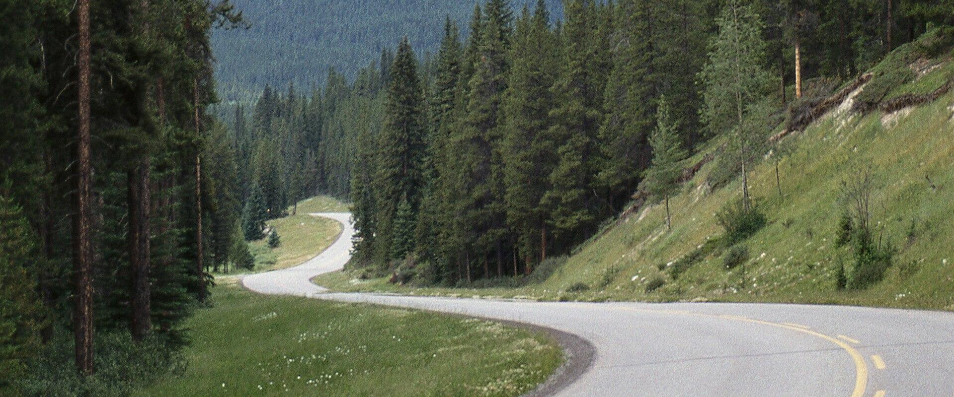 A paved two-lane road curves through a forested mountain landscape with a prominent rocky peak in the background under a cloudy blue sky.