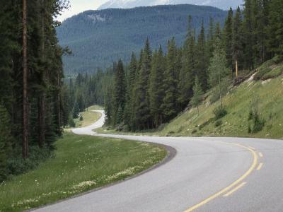 A paved two-lane road curves through a forested mountain landscape with a prominent rocky peak in the background under a cloudy blue sky.