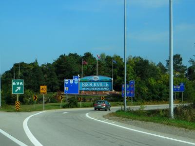 A highway interchange scene showing a large welcome sign reading "BROCKVILLE CITY OF THE 1000 ISLANDS" alongside road signs for Highway 696 and Highway 401.