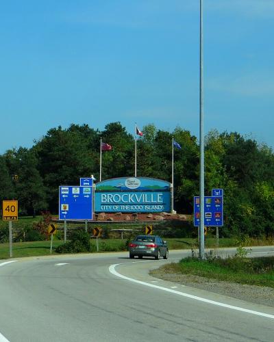 A highway interchange scene showing a large welcome sign reading "BROCKVILLE CITY OF THE 1000 ISLANDS" alongside road signs for Highway 696 and Highway 401.