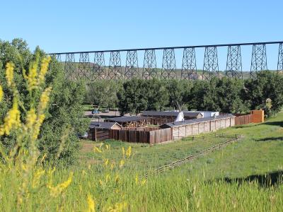A wooden palisade fort with a teepee inside, set in a grassy field with yellow wildflowers in the foreground and a large steel railway trestle bridge spanning the background.