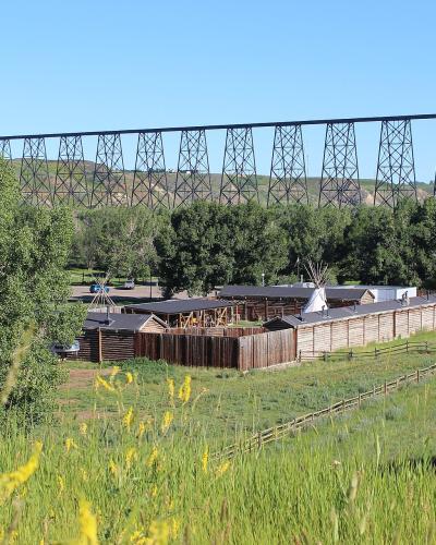 A wooden palisade fort with a teepee inside, set in a grassy field with yellow wildflowers in the foreground and a large steel railway trestle bridge spanning the background.