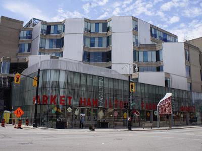 A street-level view of a modern building with a glass facade featuring large red lettering that reads "MARKET HAMILTON" and "FARM MARKET", situated at an urban intersection with traffic lights and construction signage.