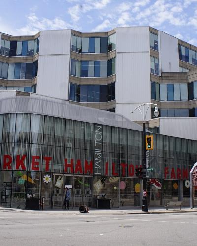 A street-level view of a modern building with a glass facade featuring large red lettering that reads "MARKET HAMILTON" and "FARM MARKET", situated at an urban intersection with traffic lights and construction signage.