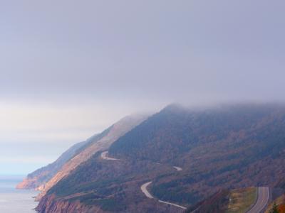 A winding coastal highway (the Cabot Trail) curves along steep, forested cliffs overlooking the ocean, with autumn foliage in the foreground and fog obscuring the mountain peaks.