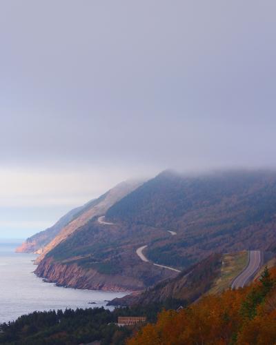 A winding coastal highway (the Cabot Trail) curves along steep, forested cliffs overlooking the ocean, with autumn foliage in the foreground and fog obscuring the mountain peaks.