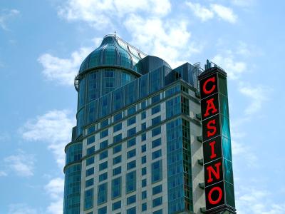 A low-angle shot of a modern high-rise hotel building with a distinctive glass-domed turret and a large vertical red neon sign reading "CASINO" attached to its side against a blue sky.