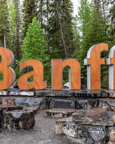 A large, wooden-lettered sign spelling "Banff" mounted on a stone base, situated beside a paved road with a forest background.