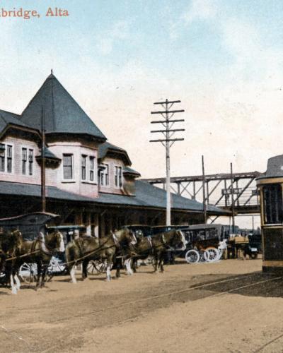 A vintage colorized postcard showing the C.P.R. Station in Lethbridge, Alberta, featuring a large brick train station building with a distinctive turret, horse-drawn carriages, and a streetcar on tracks in the foreground.