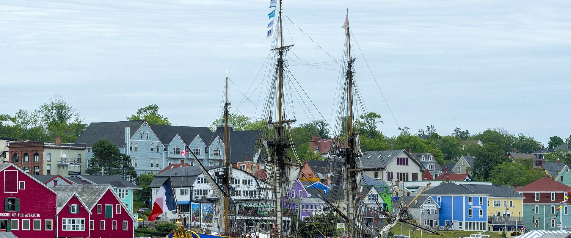 A large, three-masted replica sailing ship (the French frigate Hermione) is docked at a wooden pier in a harbor, with a backdrop of colorful, historic wooden buildings on a hillside.