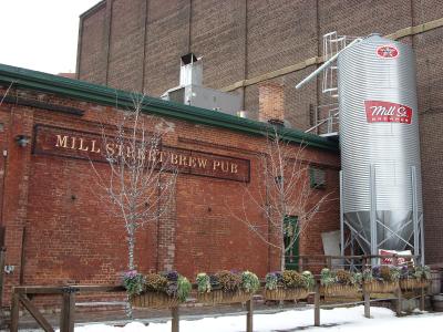 A red brick building exterior featuring a large sign reading "MILL STREET BREW PUB" and a tall silver grain silo with a matching logo, set against a snowy ground.
