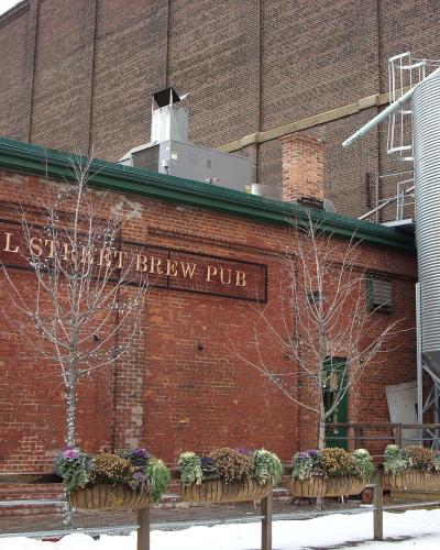 A red brick building exterior featuring a large sign reading "MILL STREET BREW PUB" and a tall silver grain silo with a matching logo, set against a snowy ground.