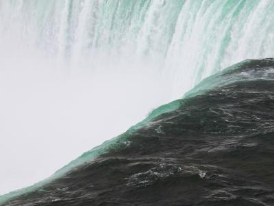 A close-up view of the crest of a large waterfall, showing the turbulent green water plunging over the edge into a cloud of white mist below.