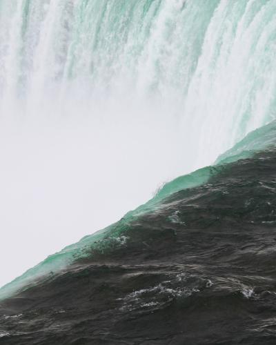 A close-up view of the crest of a large waterfall, showing the turbulent green water plunging over the edge into a cloud of white mist below.