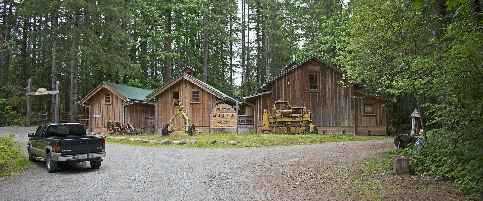 A gravel driveway leading to a cluster of wooden mill buildings surrounded by a dense forest, with a pickup truck parked on the left and a sign reading "WELCOME TO MCLEAN MILL" in the center.