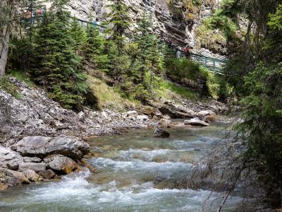 A rushing stream flows through a rocky canyon bed with steep limestone walls, while a green metal catwalk with hikers is visible along the upper left cliffside.