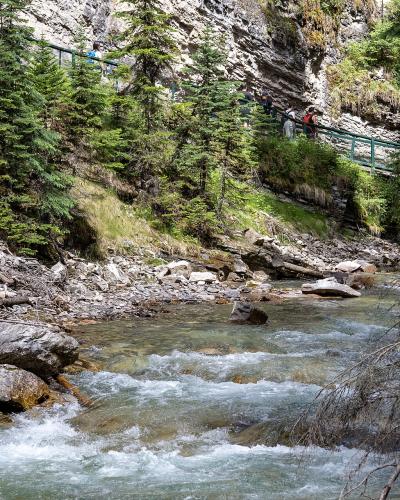 A rushing stream flows through a rocky canyon bed with steep limestone walls, while a green metal catwalk with hikers is visible along the upper left cliffside.