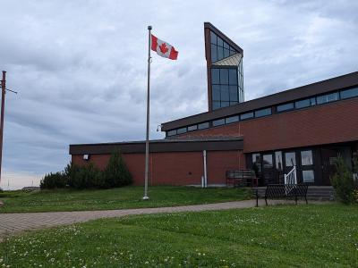 A red brick building with a distinctive tall, angular glass tower section, a Canadian flag flying on a pole, and a paved walkway leading to the entrance.