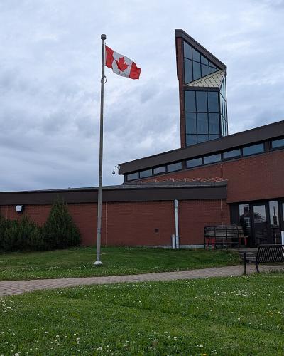 A red brick building with a distinctive tall, angular glass tower section, a Canadian flag flying on a pole, and a paved walkway leading to the entrance.