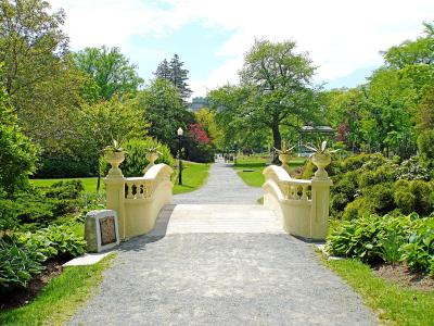 A gravel path leading across a small, cream-colored stone bridge with decorative urns, surrounded by lush green gardens and trees, with a gazebo visible in the background.