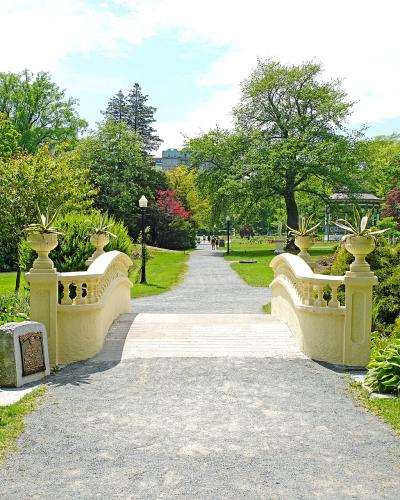 A gravel path leading across a small, cream-colored stone bridge with decorative urns, surrounded by lush green gardens and trees, with a gazebo visible in the background.