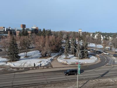 A high-angle view of a road intersection with snow on the ground, featuring a green banner on a lamppost in the foreground and university-style buildings in the background.