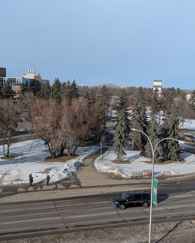 A high-angle view of a road intersection with snow on the ground, featuring a green banner on a lamppost in the foreground and university-style buildings in the background.