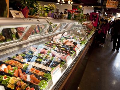 An indoor market scene showing a long refrigerated display case filled with packaged meats, sausages, and prepared foods, with shoppers walking along the aisle.